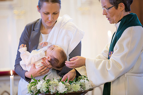 Nainen pitää sylissään vauvaa, jota pappi kastaa vedellä.